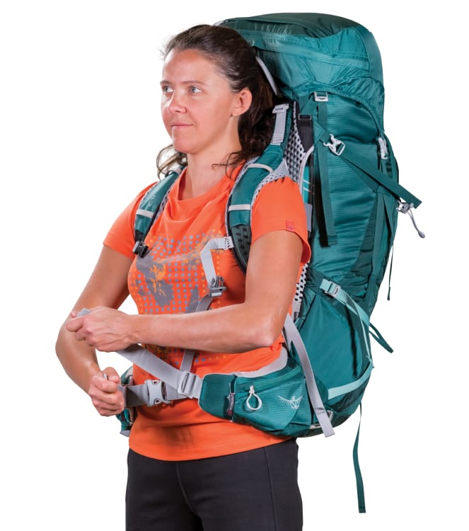 Eine Frau in einem orangefarbenen T-Shirt und einem blaugrünen Rucksack über den Schultern zieht den Nylon-Verstellgurt des Hüftgurts fest.