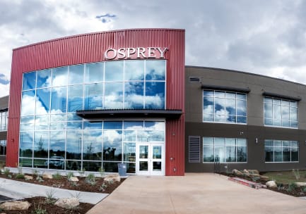 Das Fischaugenfoto eines großen, anthrazitfarbenen und roten, rechteckigen Bürogebäudes mit einer Fensterscheibe an der Hauptfassade und einem Schriftzug aus Edelstahl mit der Aufschrift „OSPREY“