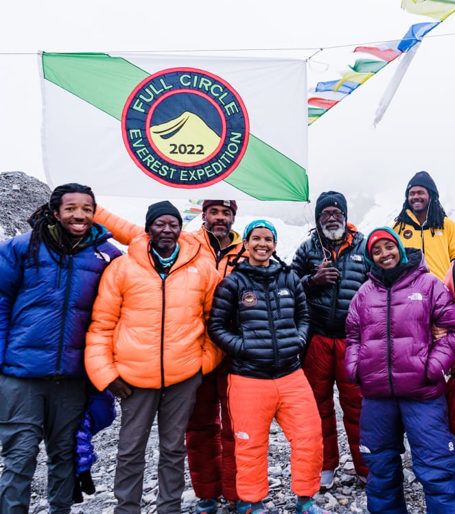 Ein Team von sieben Bergsteigern in Bergsteigerhosen und dicken Mänteln unter einer weiß-grün gestreiften Flagge mit blauen, roten und gelben Abzeichen, auf denen „Full Circle Everest Expedition 2022“ steht, und tibetischen Gebetsfahnen, die hinter ihnen im Wind wehen.