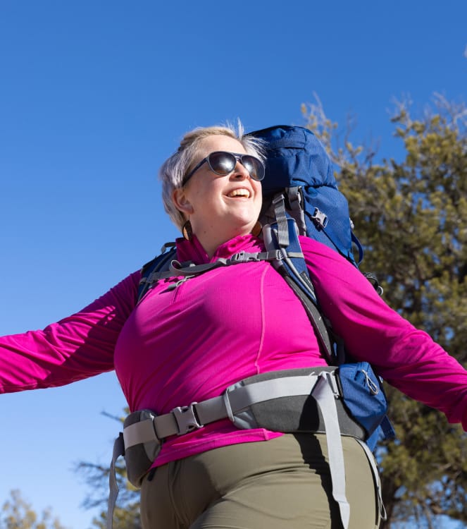 Ein Foto von einer Frau mit silbernen Haaren, die einen Fuchsia-farbenen Pullover mit halbem Reißverschluss, eine Wanderhose und einen kobaltblauen und grauen Osprey-Rucksack unter einem blauen Himmel trägt.