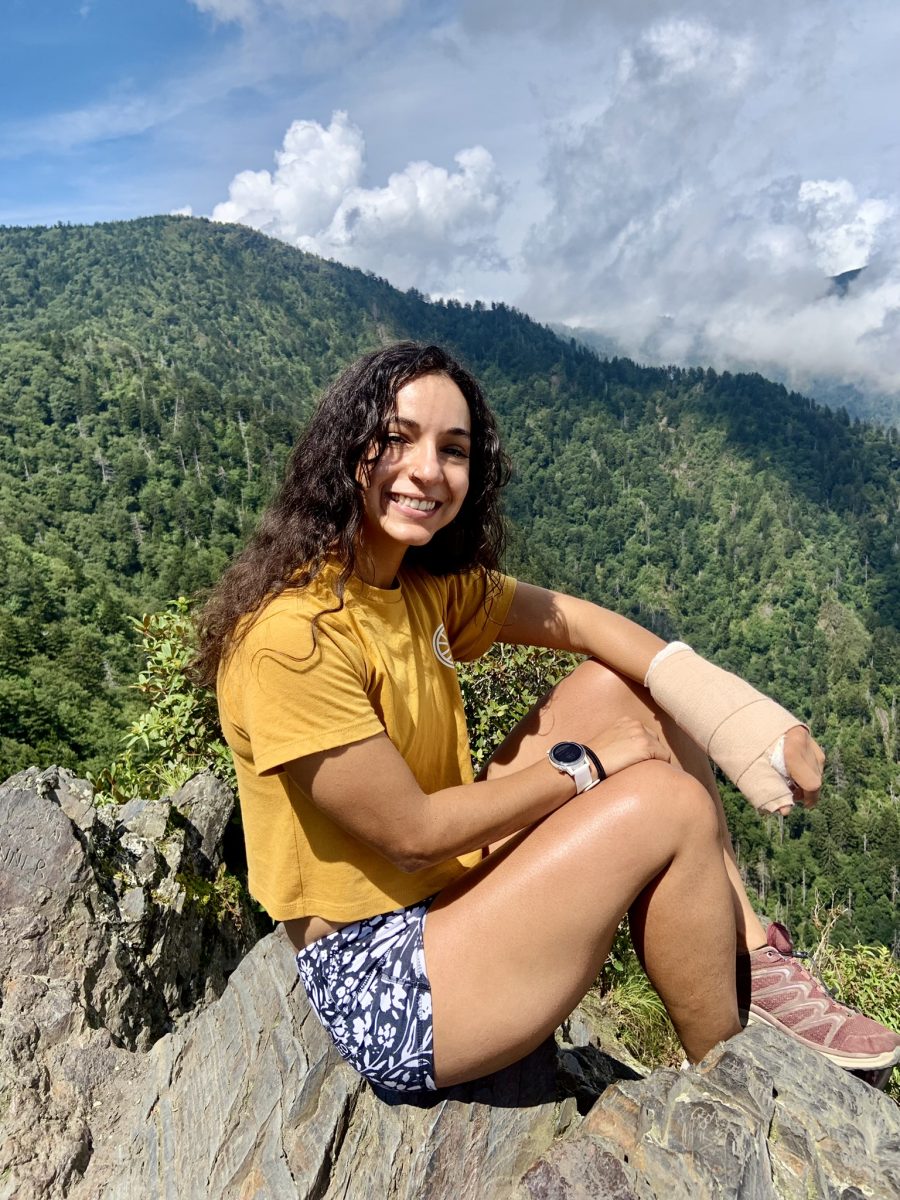 A woman wearing a yellow shirt and blue shorts sitting atop a rocky mountain overlook, with a soft cast on her left arm
