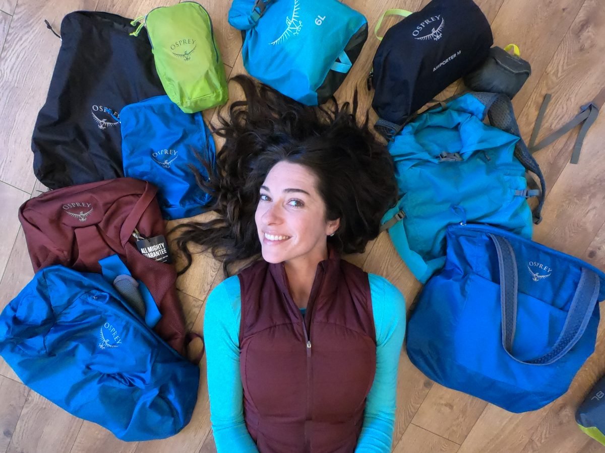 A woman wearing a teal shirt and a maroon vest laying on the ground surrounded by travel accessories
