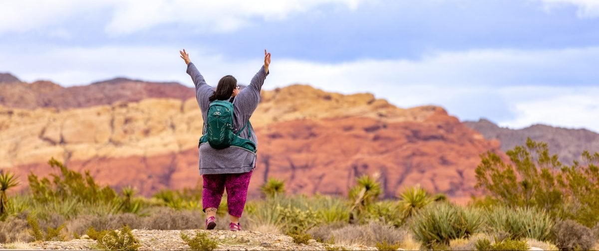 A woman facing in the Nevada desert wearing a teal backpack and holding her hands in the air