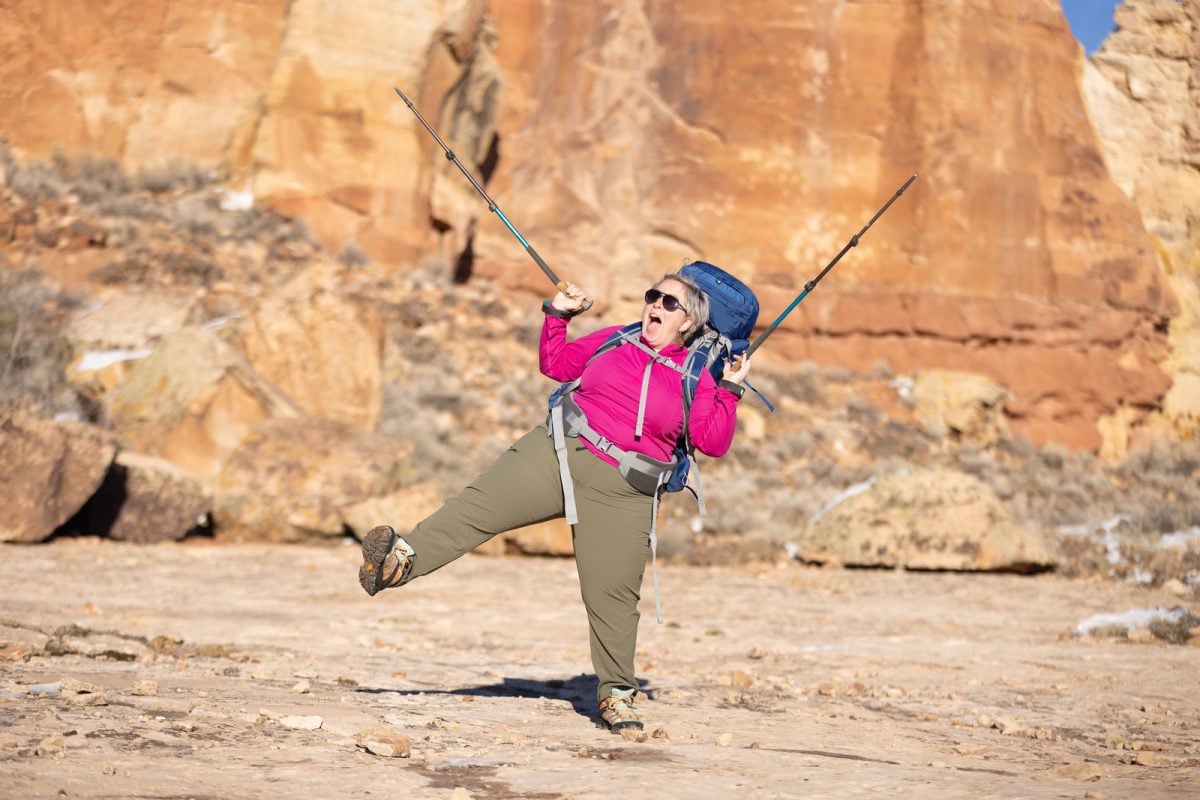 A woman wearing backpacking gear holding her hiking poles up in celebration while lifting one leg off the ground