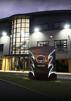A nighttime photo of a 3-story office building with a curved roof and a multi-story glass atrium at the front entrance. There is a large, illuminated sign on a patch of grass in front of the building, featuring the Osprey logo, wing-like cutouts that are lit up, and the name &ldquo;Talon House.&rdquo;