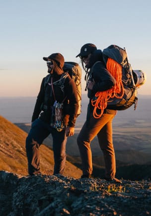 Two climbers are standing in morning light on a mountain ridge, wearing fully-loaded packs with a climbing rope, carabiners, helmets and an ice ax.