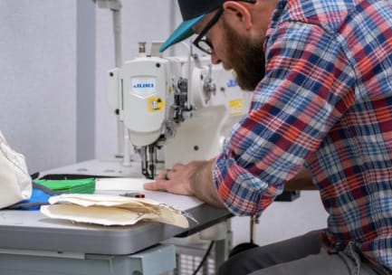 A bearded man wearing glasses, a baseball cap and a blue, white and red flannel shirt with rolled up sleeves constructs a cloth face mask on a sewing machine.